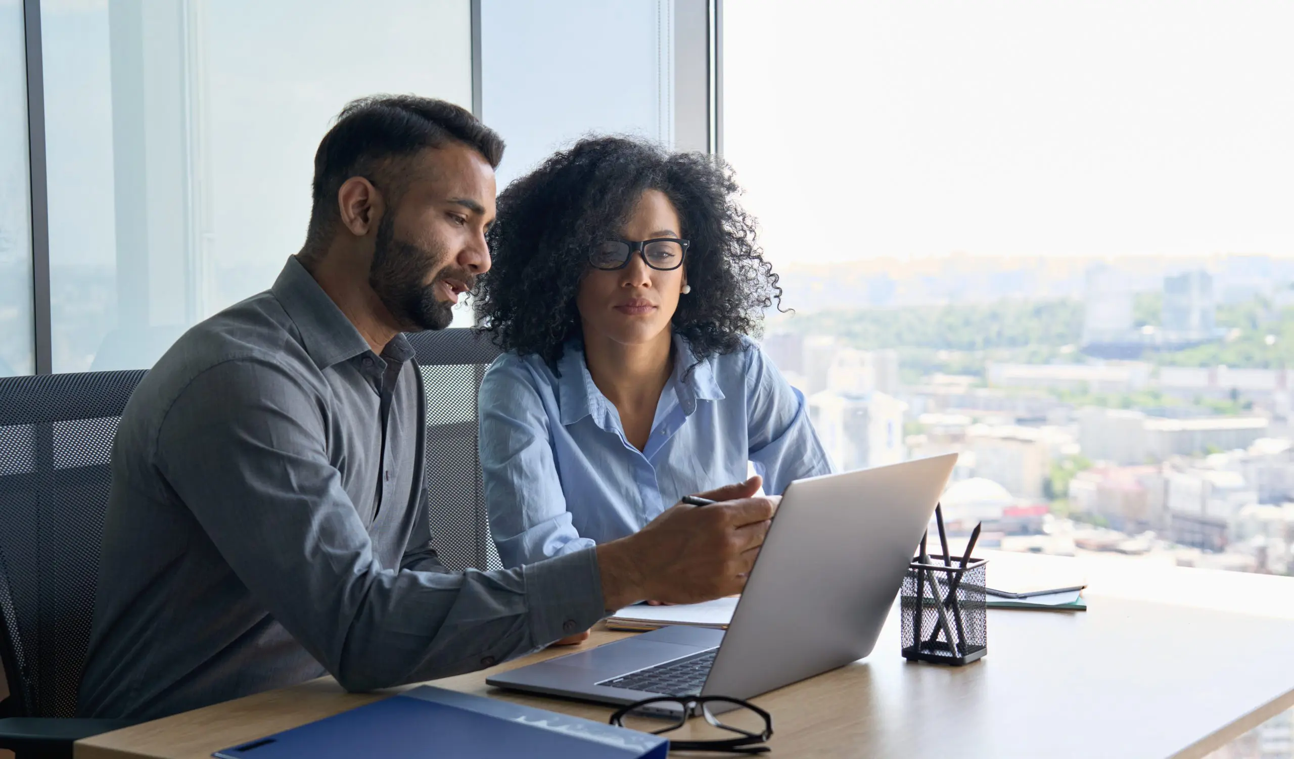 Two colleagues sit at a desk in an office, collaborating while looking at a laptop screen.