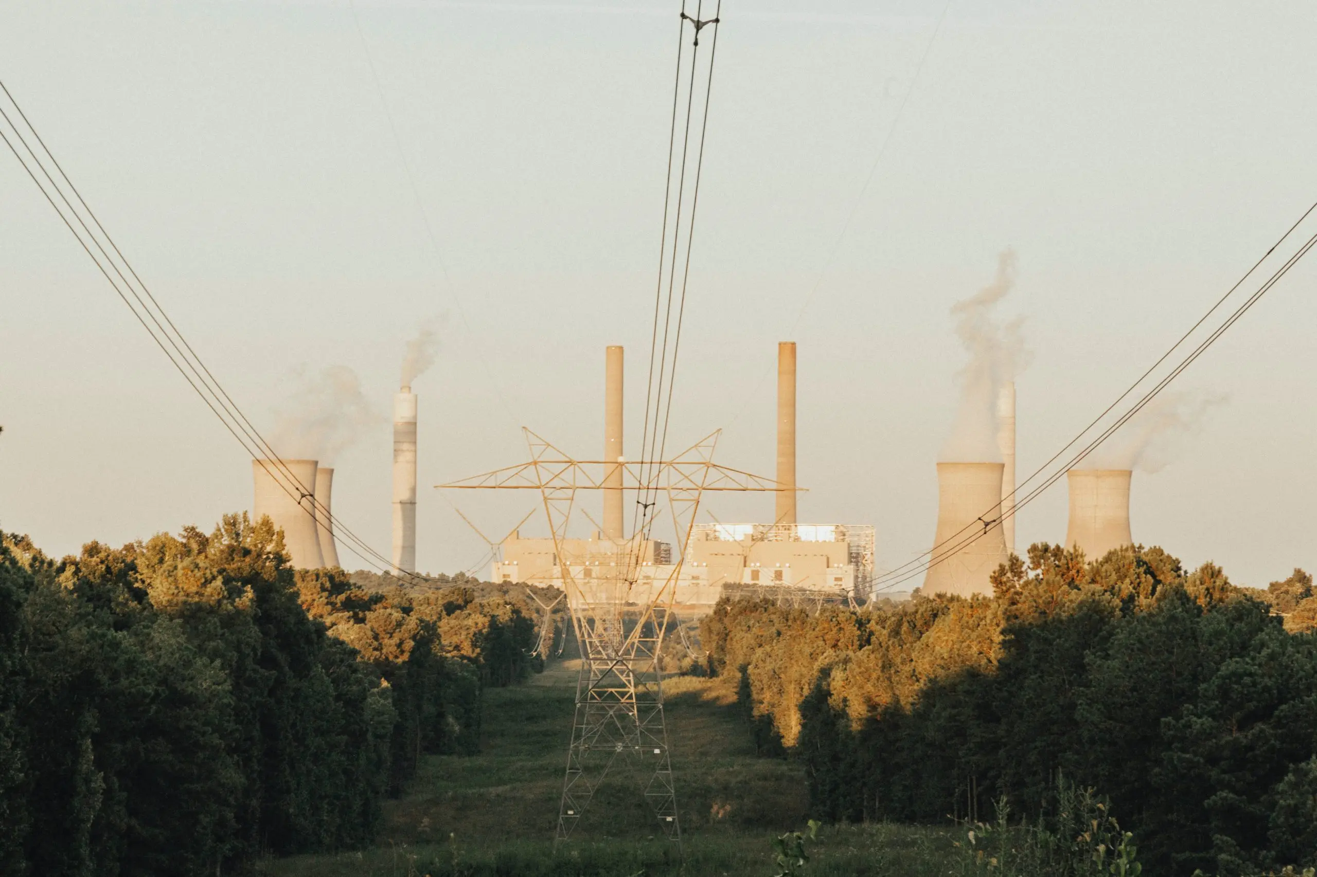 Power lines lead to a large industrial plant with smokestacks emitting smoke, surrounded by a dense forest.