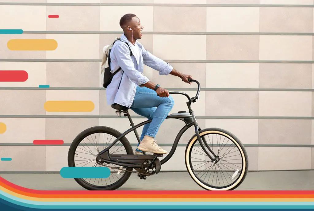 A smiling male cyclist wearing casual clothes is riding a bike with wired earbuds in his ears and a backpack on his back.