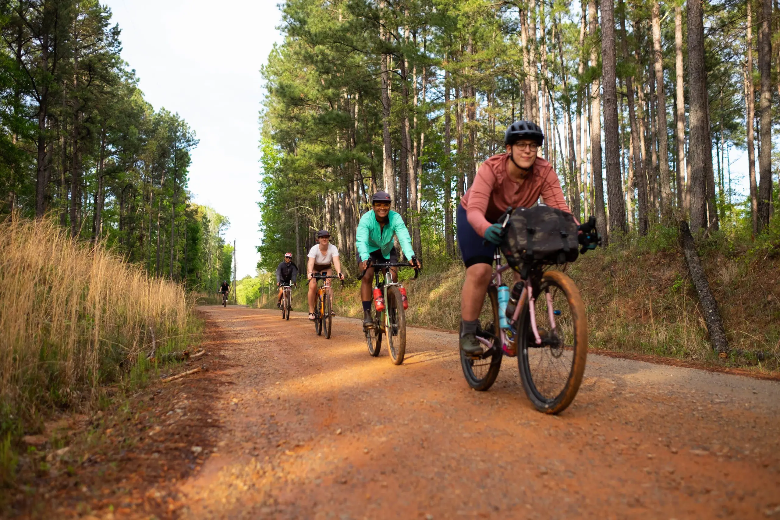 Five people are wearing helmets and riding bikes down a clay path with grass and tall trees on either side.