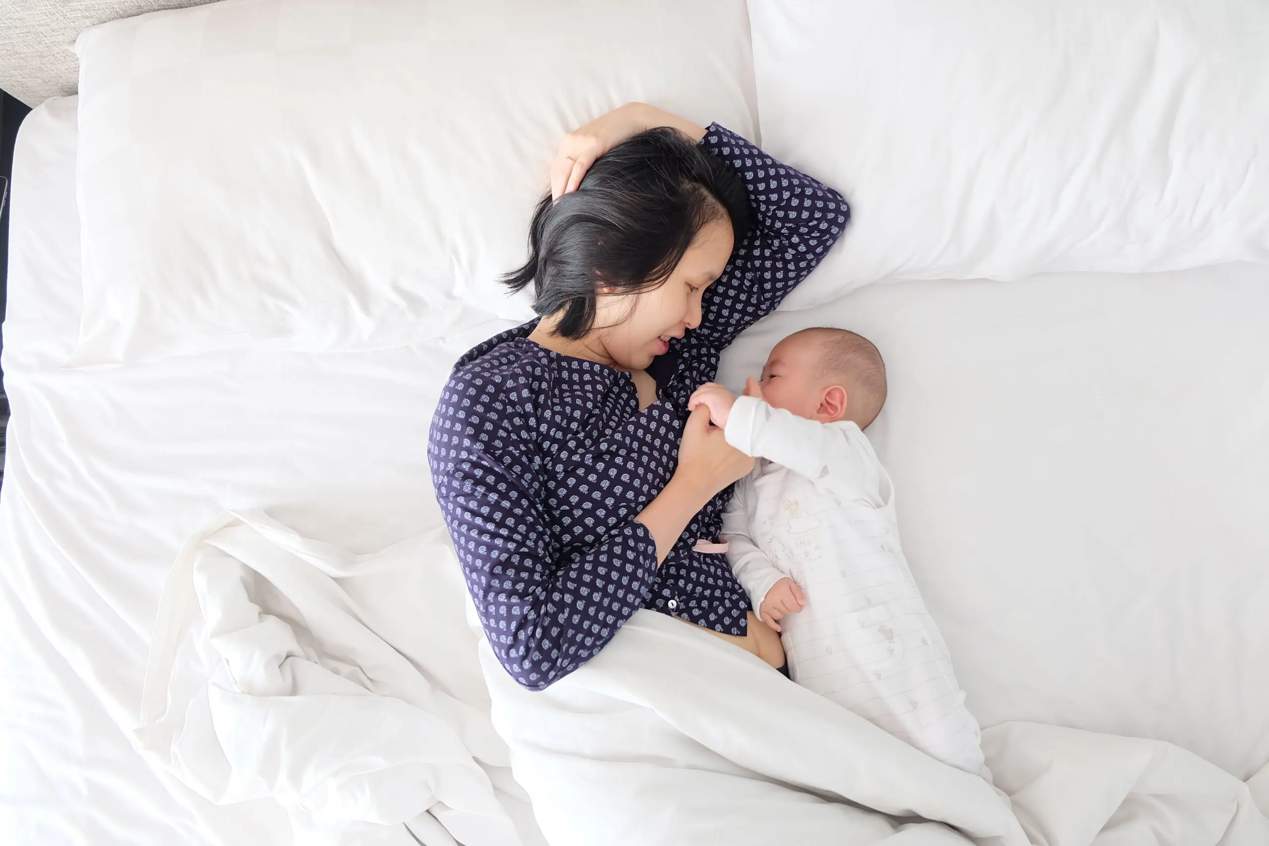 A woman wearing a navy blue blouse is laying in a bed with her left hand behind her head. Next to her is an infant dressed in a white onesie who is holding onto the woman's right thumb.