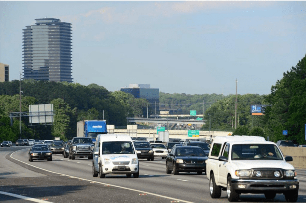 Several cars are driving on a freeway including black sedans, a white truck, a black truck, and a white van.