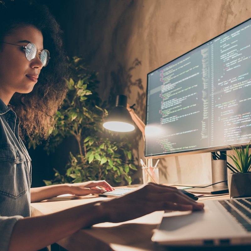 A young woman wearing glasses is focused intently on her work while using a computer and monitor at her desk.