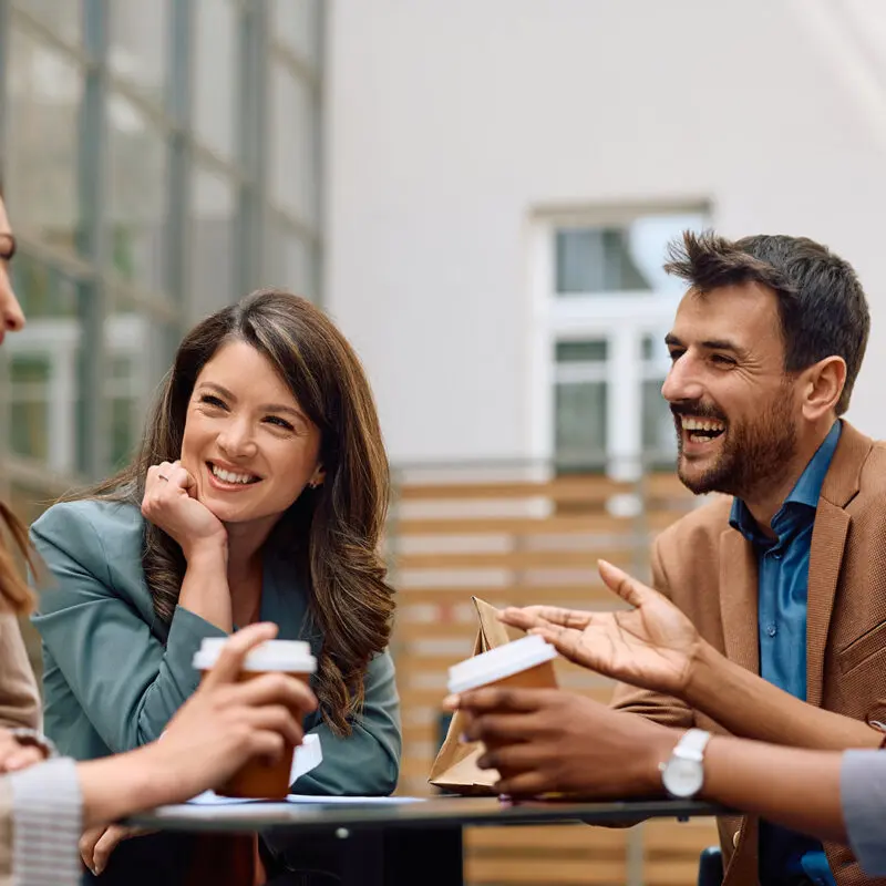 A group of four people are sitting outdoors, smiling and chatting over coffee in a relaxed, professional setting.