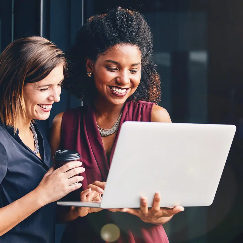 Two women are smiling and looking at a laptop that one of them is holding. The other woman looks on with a coffee cup in her right hand.