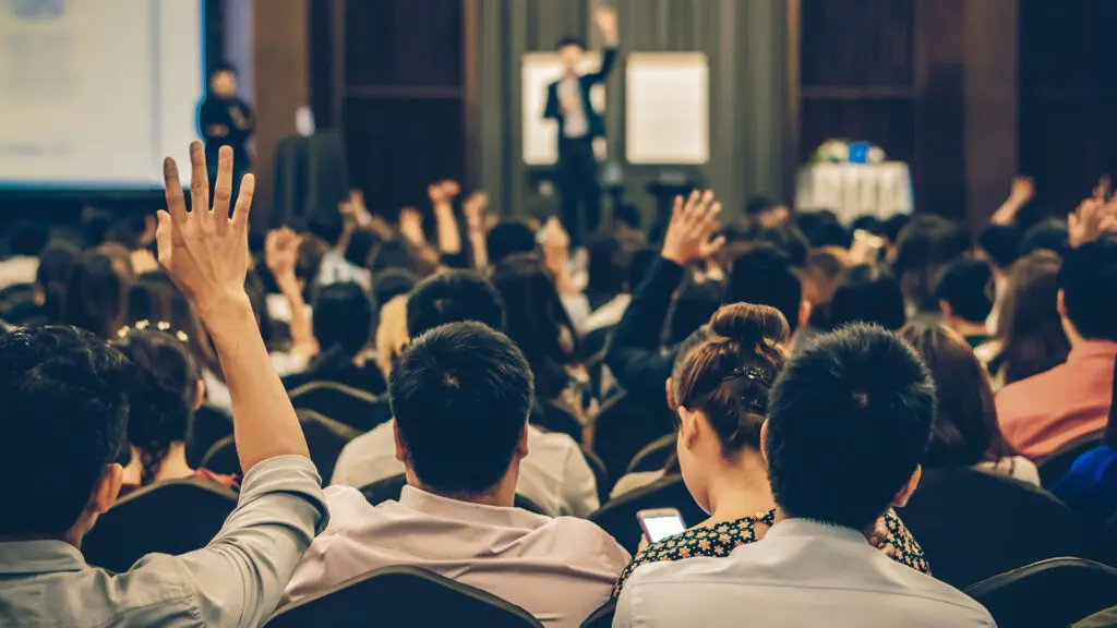 People are seated shoulder to shoulder in a crowded conference room. They face forward with their backs to the camera. The speaker and several people seated in the room have their hands raised.