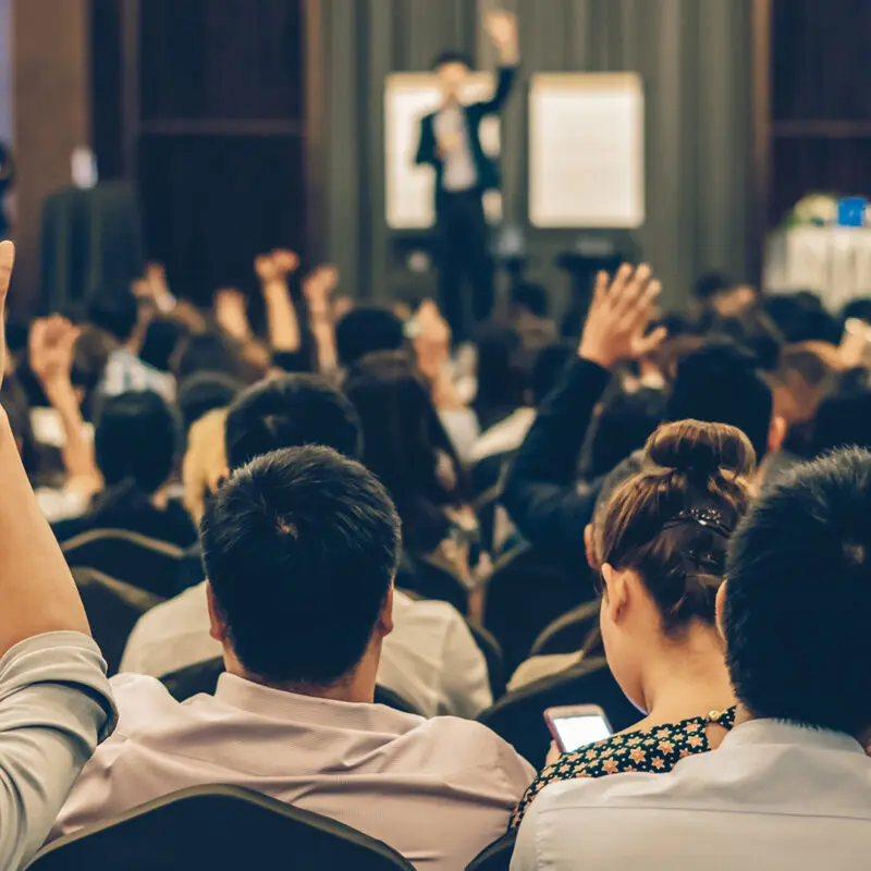 People are seated shoulder to shoulder in a crowded conference room. They face forward with their backs to the camera. The speaker and several people seated in the room have their hands raised.