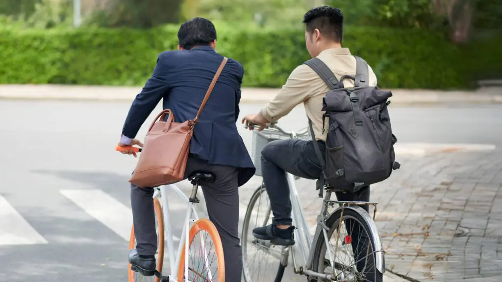 Back view of two people wearing bags while riding bicycles on a path with greenery.