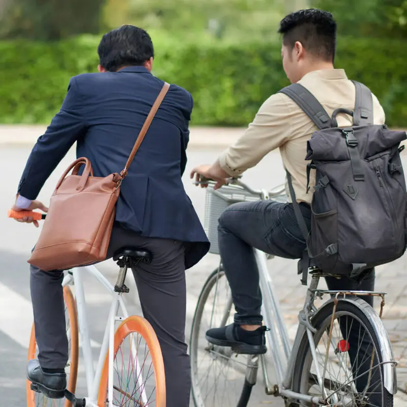 Back view of two people wearing bags while riding bicycles on a path with greenery.