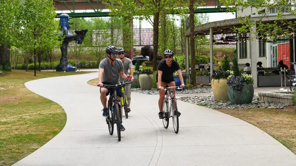 Three people wearing helmets ride bicycles on a curving path through a park with trees, plants, and a cafe nearby. Other people are seated outside the cafe. The scene is bright and lively.