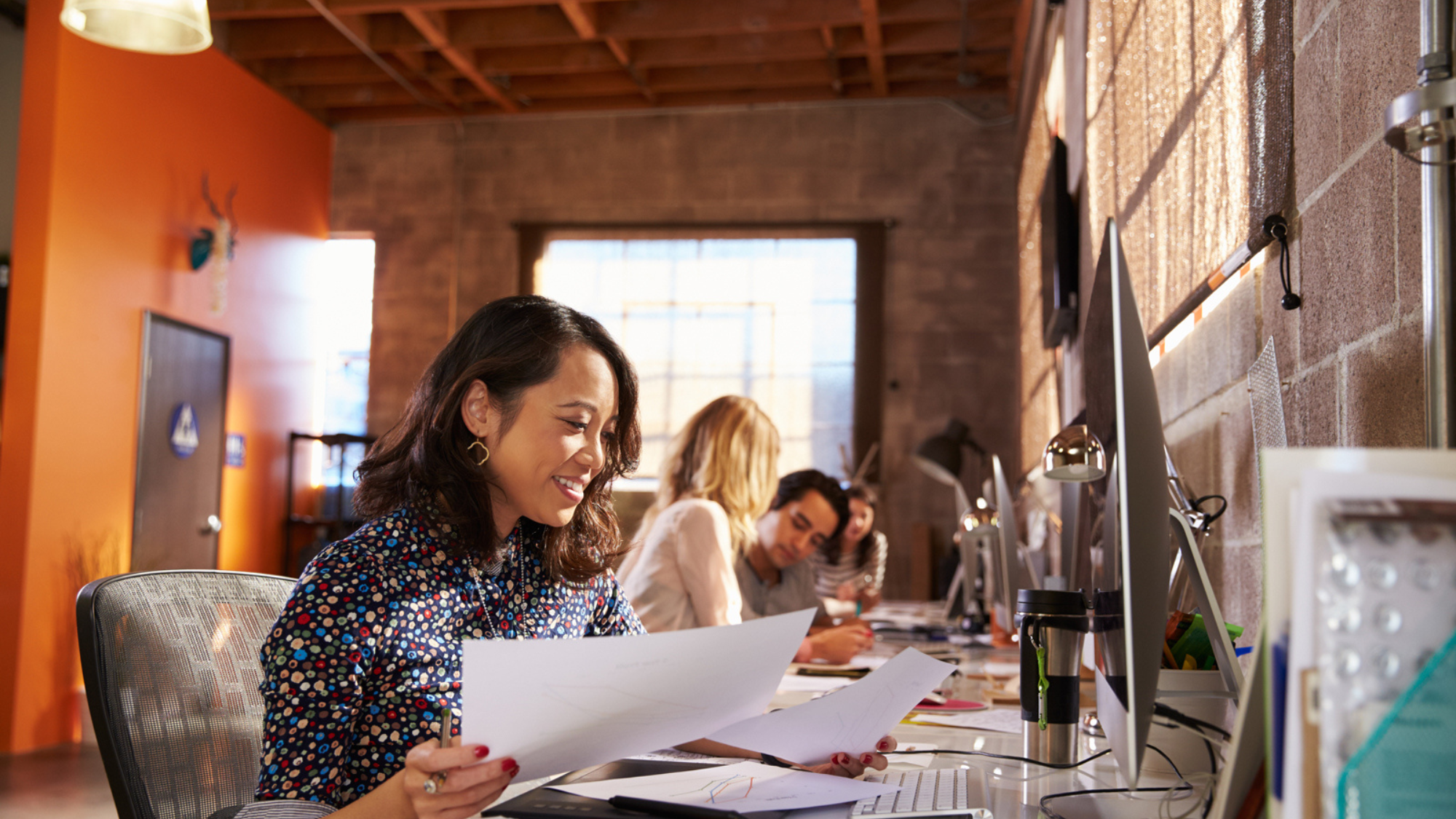 A woman is smiling and looking at two documents in her hand while sitting at a table with a computer in front of her. Three other people are seated to the left of her with computers in front of them as well.