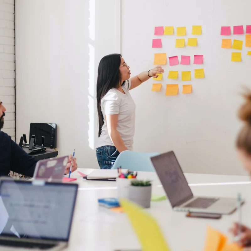 Five people are in a meeting together. One woman is speaking at the front while looking at several sticky notes on the wall.