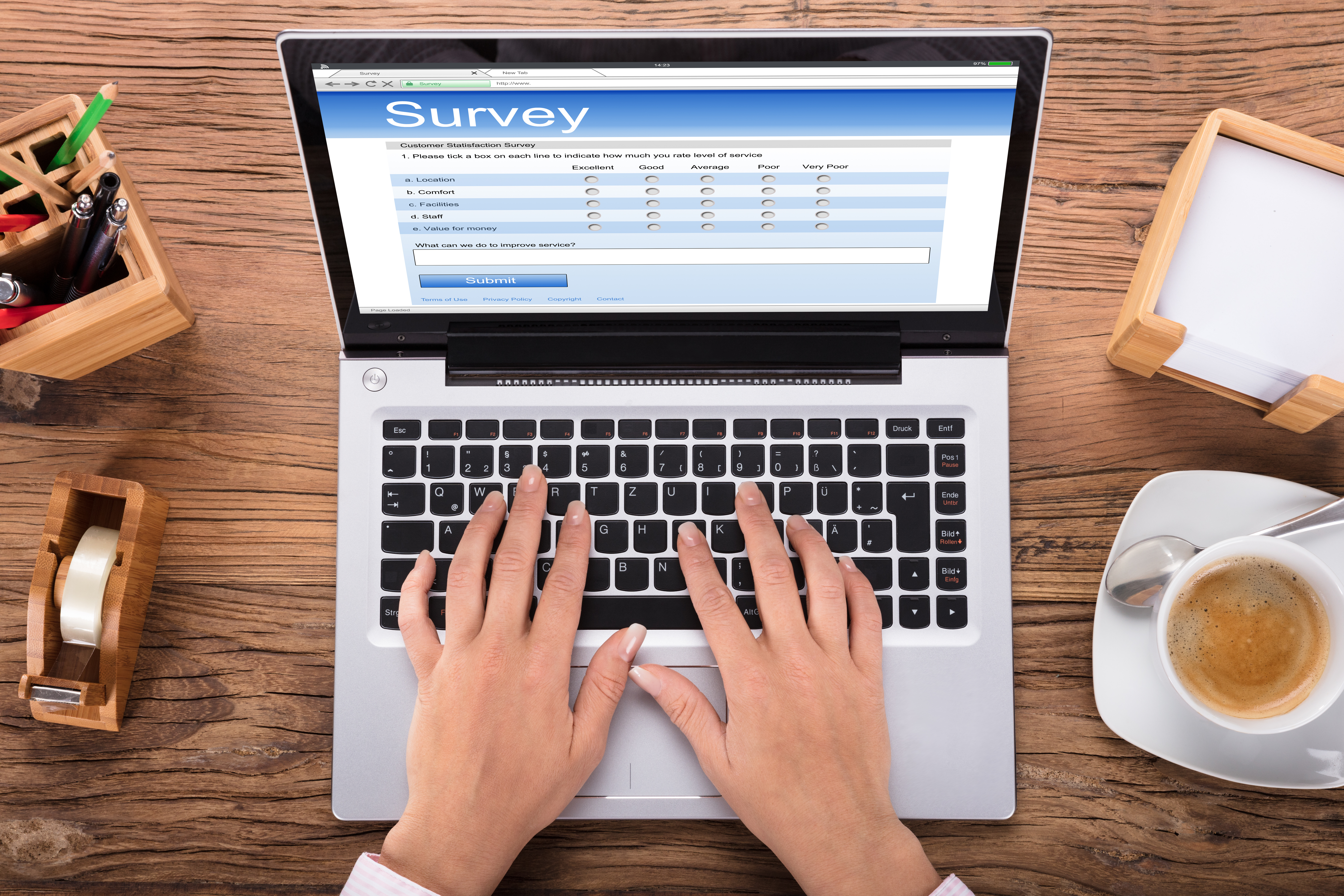 Elevated view of a woman's hands doing a survey on a laptop. On the table, along with the laptop, is a tape dispenser, penholder, coffee mug and coaster, and a stack of square papers.