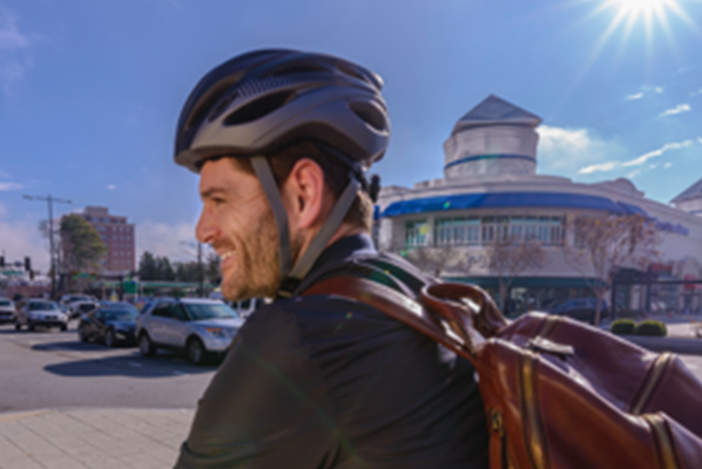 A man is smiling and wearing a bike helmet and brown backpack. There are several cars and a building in the background.
