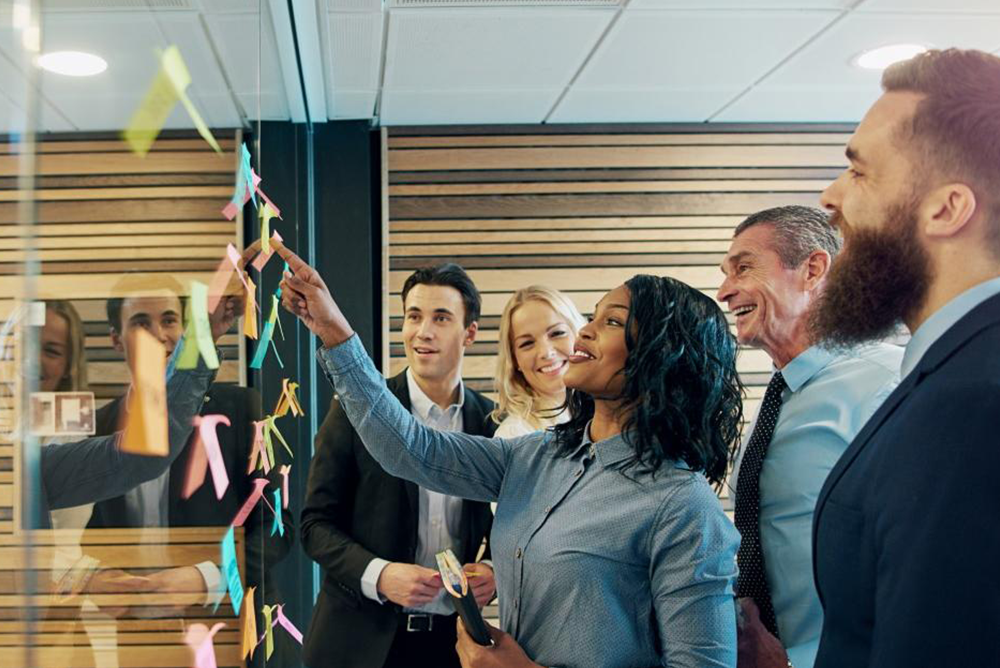 Two women and three men dressed in business casual clothes are gathered around a glass wall with multiple post-its on it. One of the women is at the front of the group pointing at a yellow post-it.