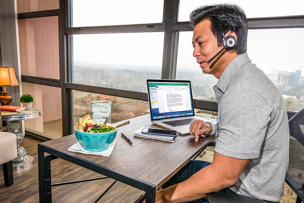 A man wearing a headset and grey shirt is sitting at a table. On the table is a laptop, notebook, smartphone, pen, cup of water, and bowl of food.