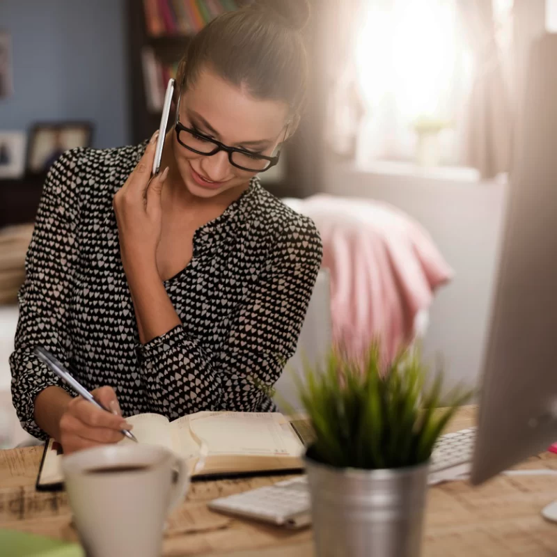 A woman wearing glasses and her hair in a bun is sitting at a desk with a computer. She is on the phone while writing something in a planner.