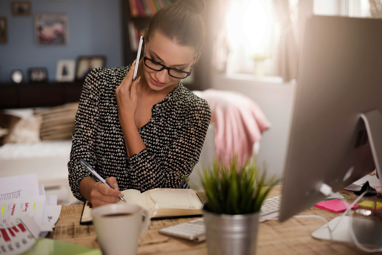 A woman wearing glasses and her hair in a bun is sitting at a desk with a computer. She is on the phone while writing something in a planner.
