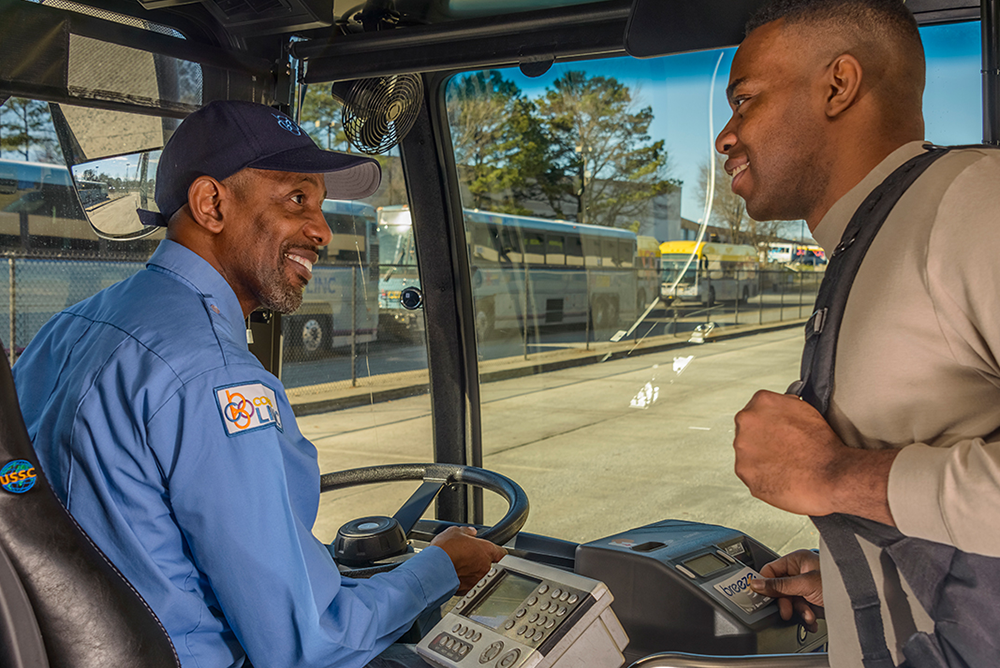 A man with a backpack over his shoulder is smiling and scanning his Breeze card. The bus driver wearing a blue shirt and hat is smiling at the man scanning his card.