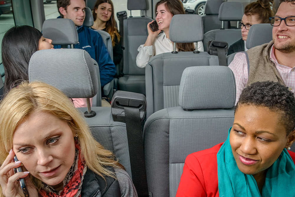 Eight commuters are in a van. Some are engaged in conversation with each other and some are talking on their phones.