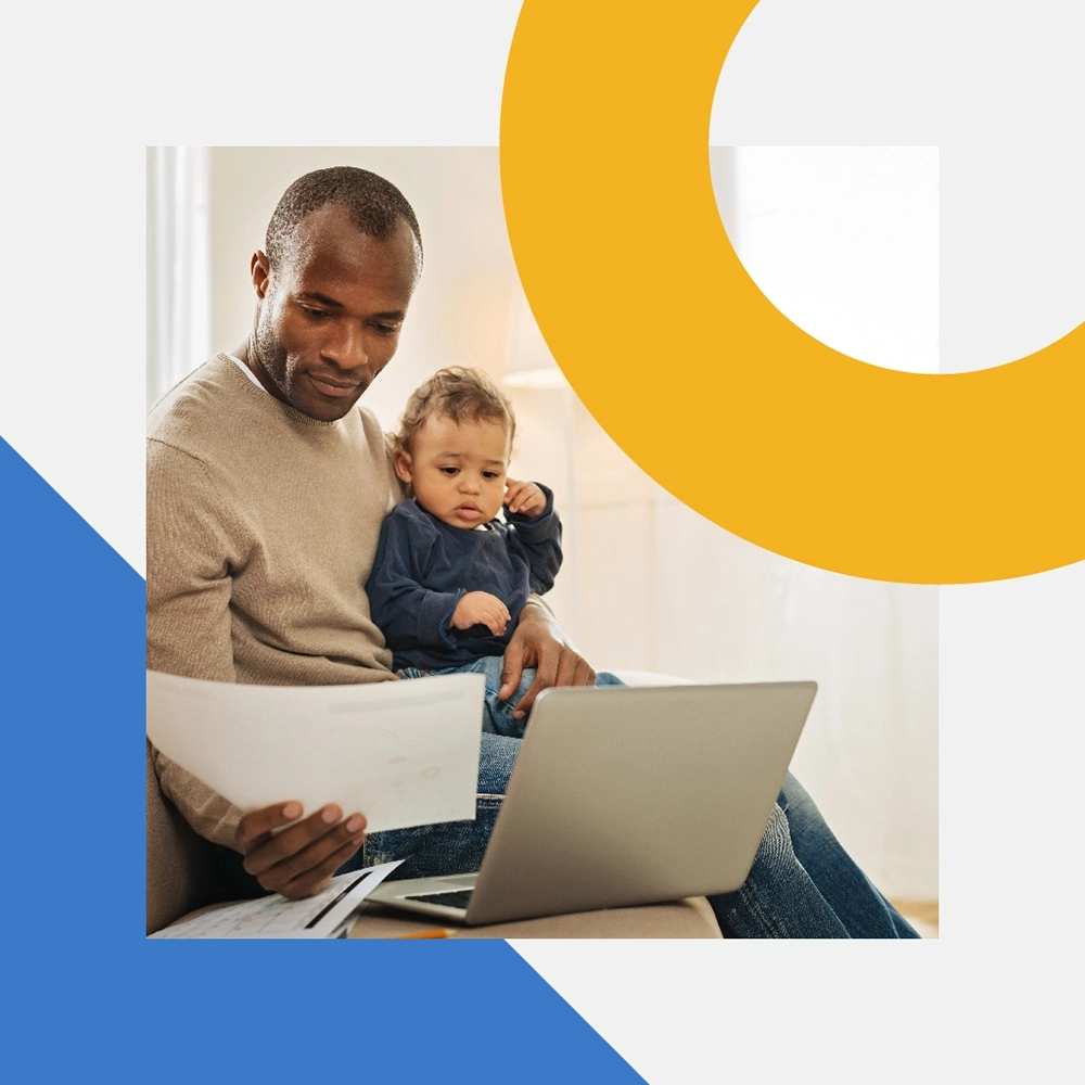 A man is holding a baby while examining a document in his right hand. There is also a laptop in front of him.