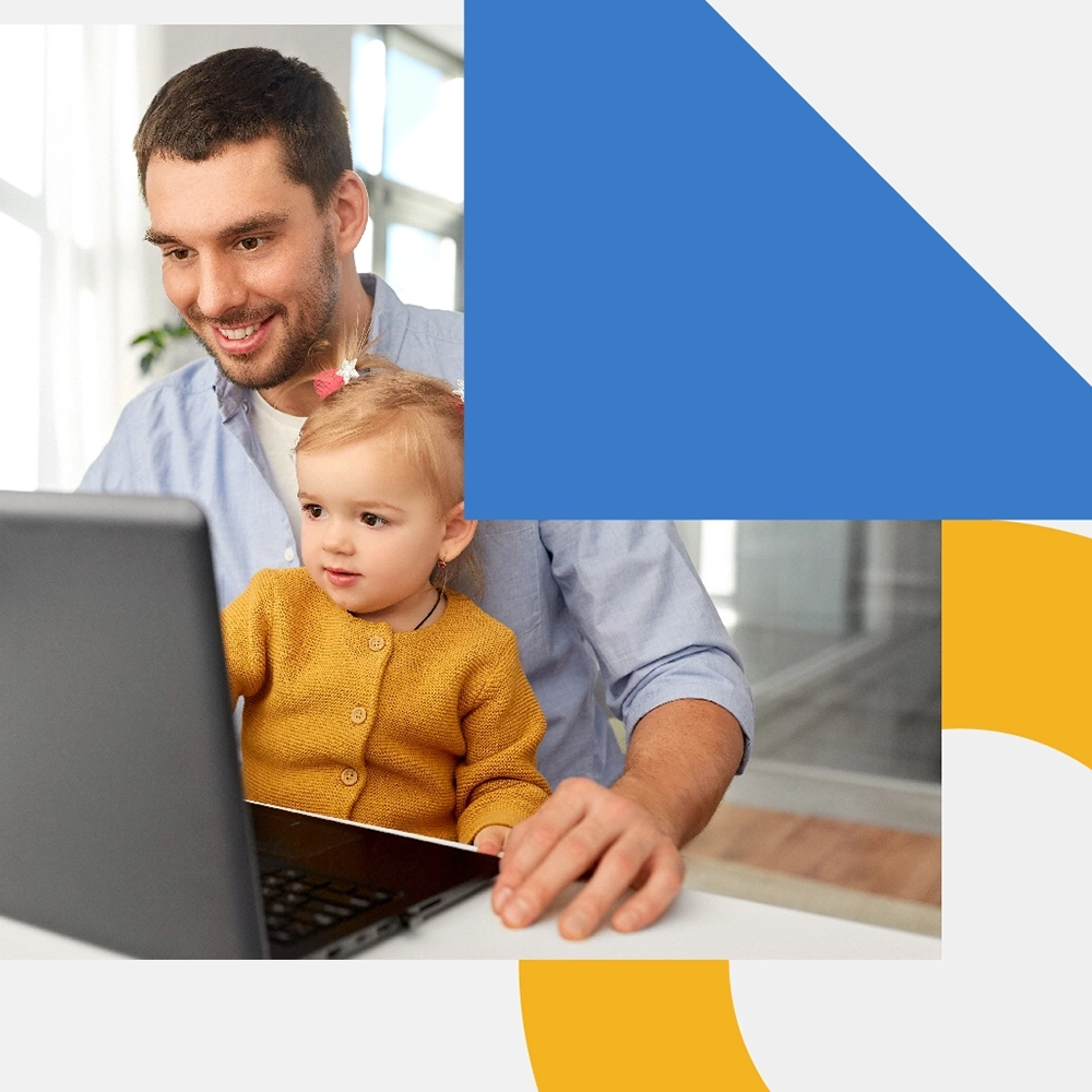 A man is sitting down and smiling while looking at the laptop on the table in front of him. There is a baby on his lap wearing a yellow sweater.