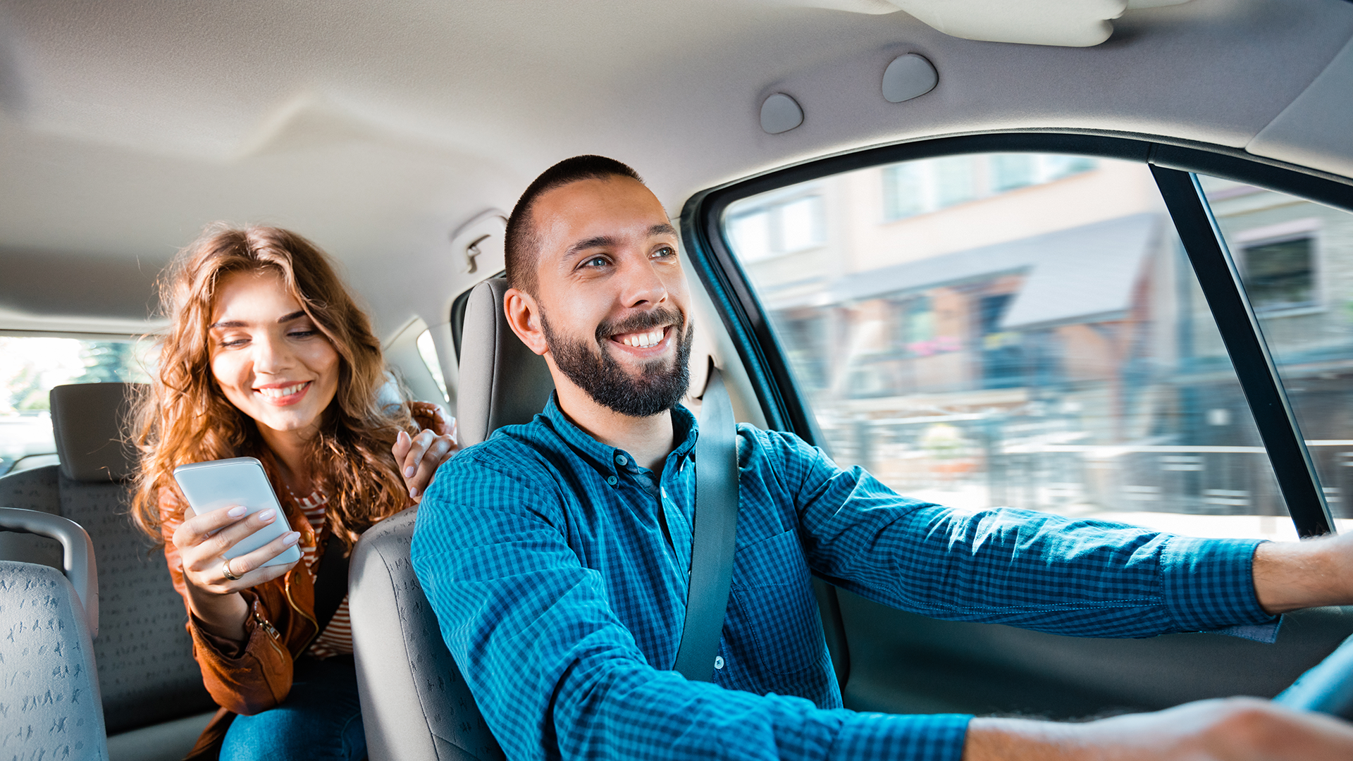 A man is smiling while driving a car. A female passenger is in the backseat smiling and looking at her phone.