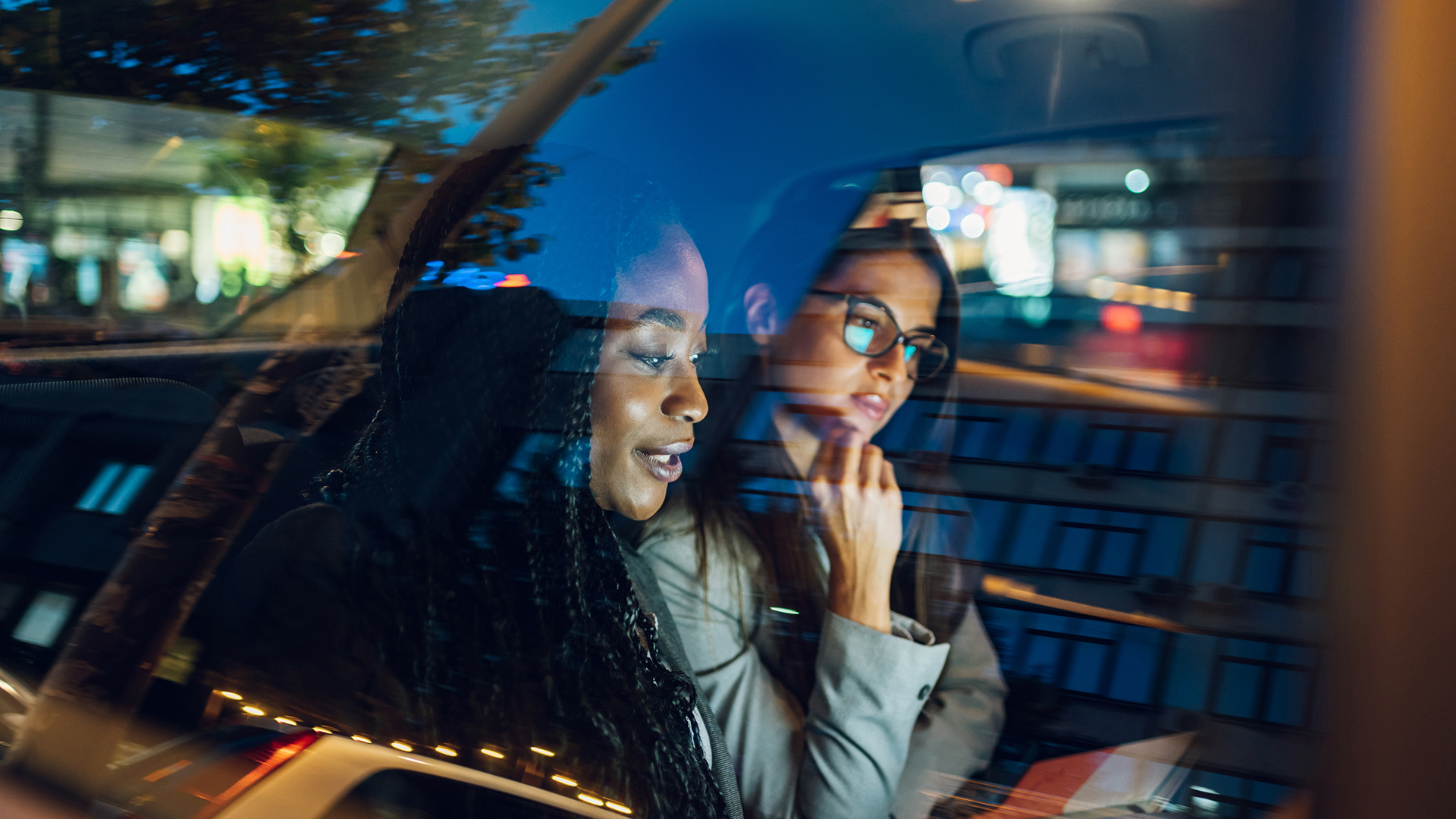 Two women are sitting in the back of a car looking at the screen of a digital device. The women on the left appears to be talking and the woman on the right has her hand on her chin.