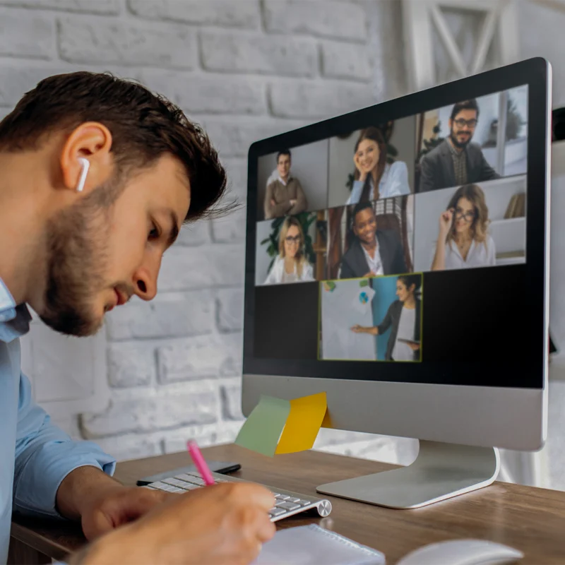 A man is writing at his desk with an earbud in his right ear. He appears to be having a meeting with seven people on the computer screen in front of him.