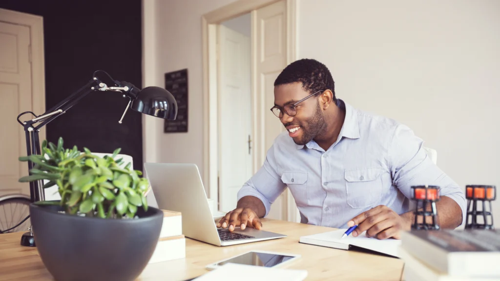 Man wearing glasses is smiling at his desk. One hand is on his laptop while the other hand is pointing at something in a book.