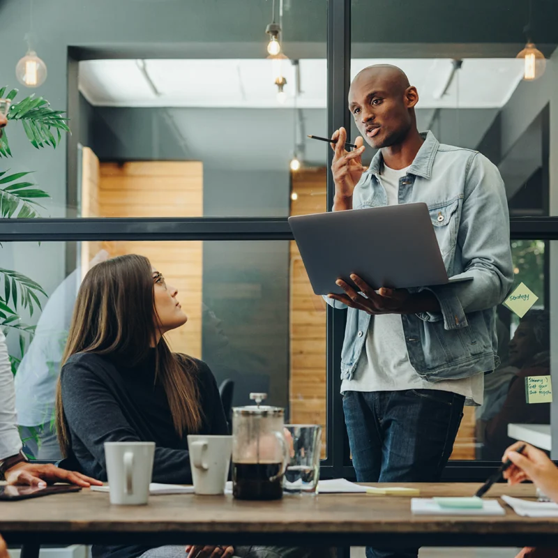 Six people are having a meeting. A man standing and holding a laptop appears to be leading their discussion.