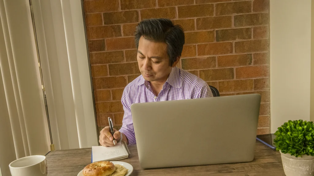 Man sits at a table while writing in a notepad. On the table is a laptop, smartphone, mug, a plate with a bagel, and the notepad.