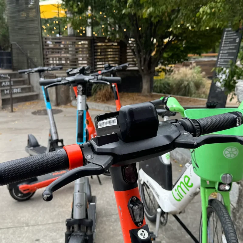 Up close of e-scooter handles with other scooters and e-bike on the sidewalk.