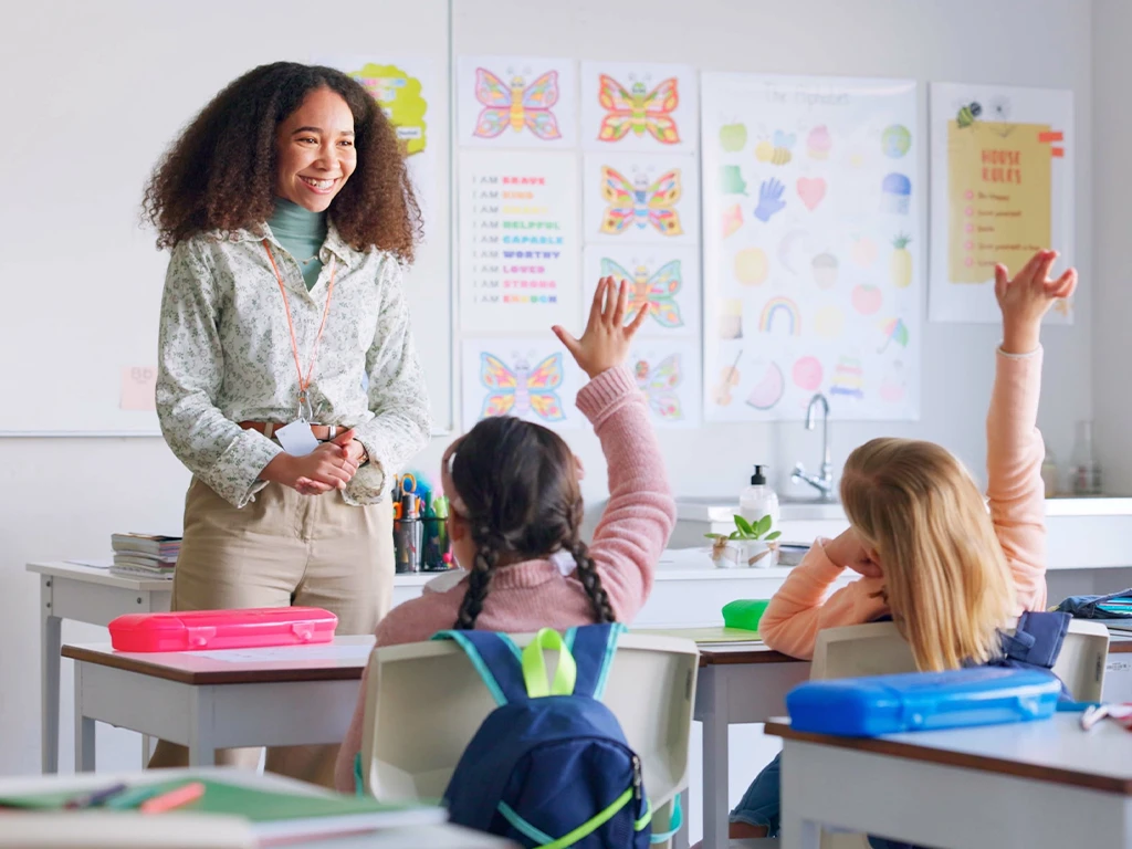 Smiling teacher at the front of a classroom with two students raising their hands.