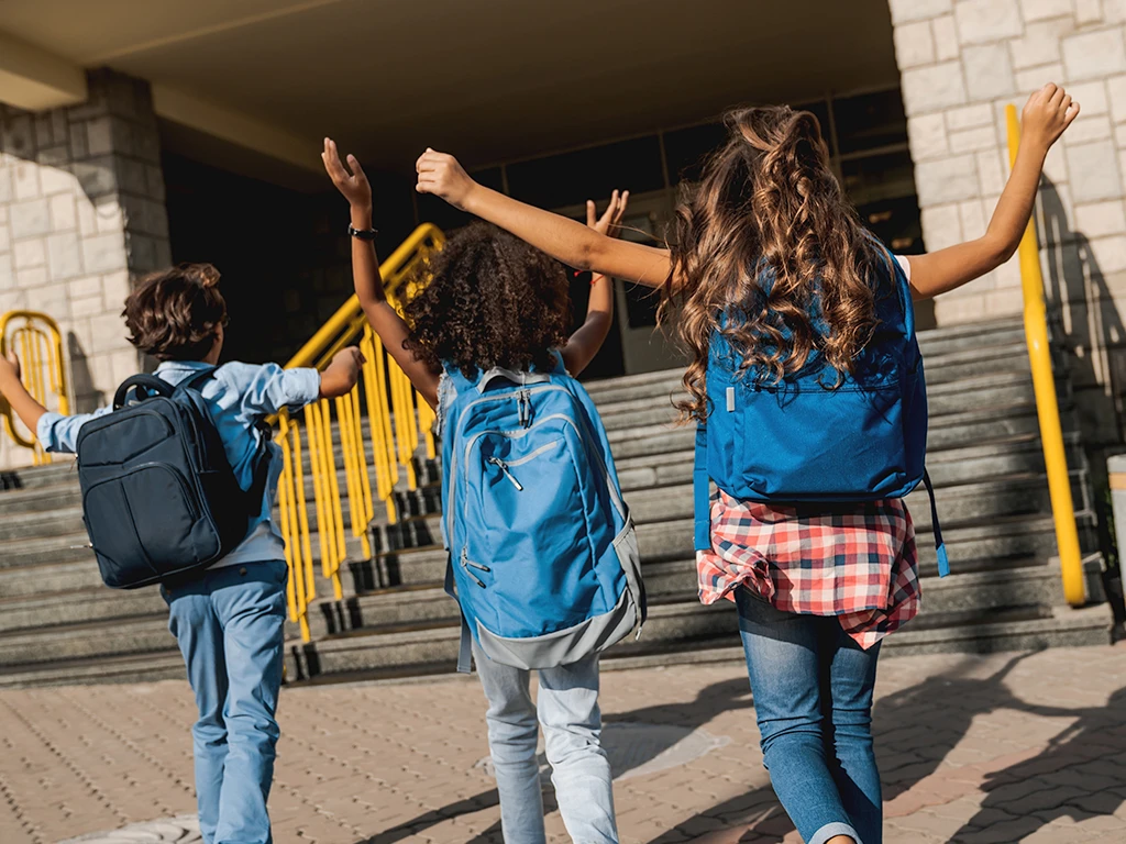 Three children excitedly walking into school with hands in the air.