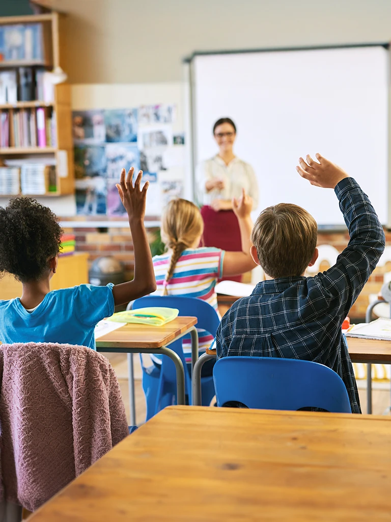 View from back of a classroom with teacher at the front and multiple children at desks with hands raised.