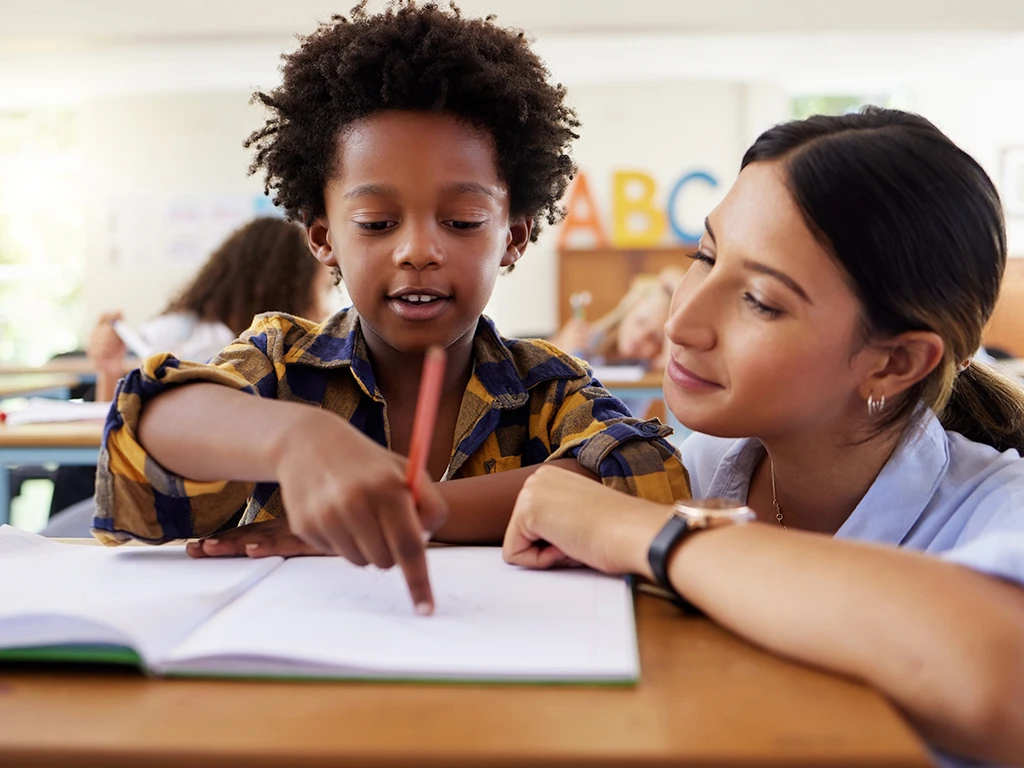 Close up of a teacher crouching down at a students desk as the student points to a notebook page.