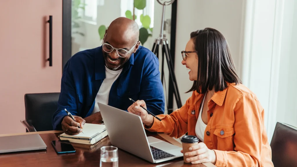 Two people working at a table and laughing. One person is writing in a notepad and the other is working at a laptop.