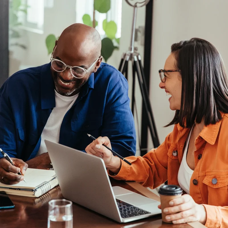 Two people working at a table and laughing. One person is writing in a notepad and the other is working at a laptop.