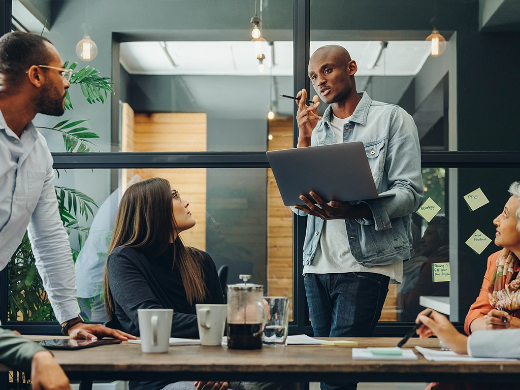 Four people are working together, one person is holding a laptop the others are listening to the person with the laptop speaking.