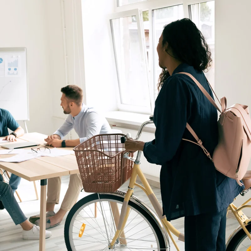 A woman to the right is wearing a backpack and holding a bicycle with a basket on the front. Two men and a woman sit around a table. The men are talking. The seated woman is waving to the woman holding the bicycle.