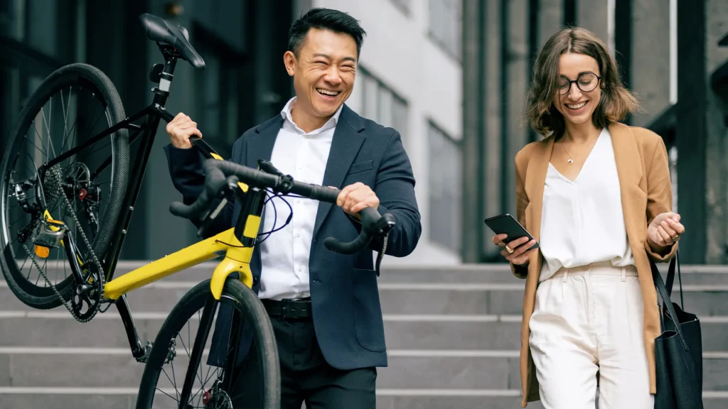 A man and a woman are walking downstairs outside of a building. The man is dressed in a suit and carrying a yellow bicycle. The woman is wearing glasses and holds a cell phone and a purse.