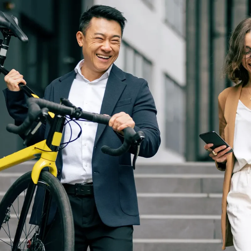 A man and a woman are walking downstairs outside of a building. The man is dressed in a suit and carrying a yellow bicycle. The woman is wearing glasses and holds a cell phone and a purse.
