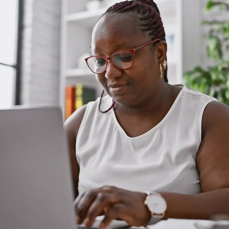 A woman wearing glasses is seated at a desk. She is looking at an open laptop computer and is typing.