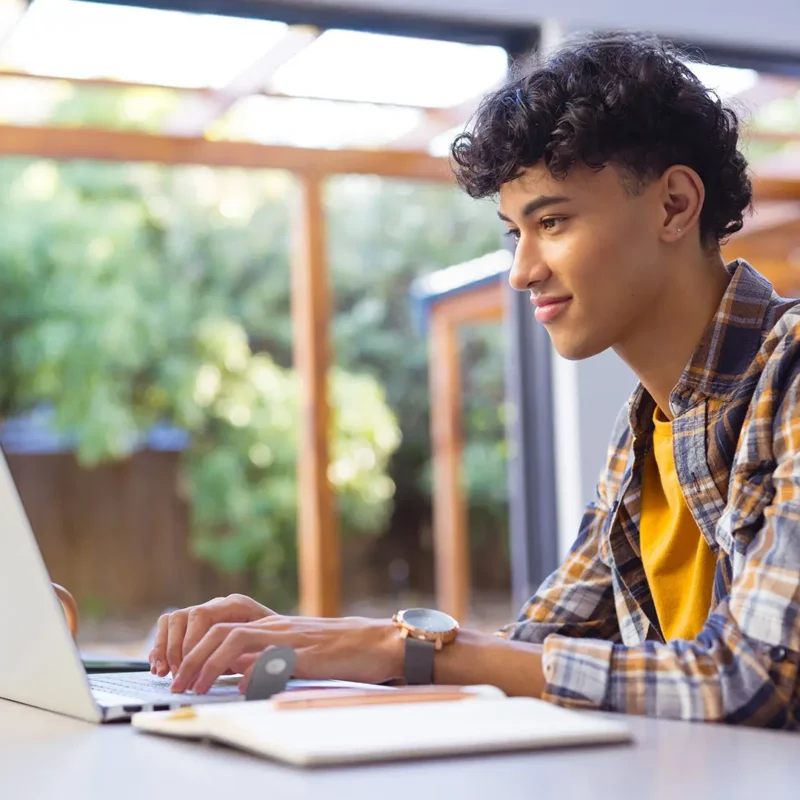 A smiling young man is using a laptop while sitting at a table in a bright setting with greenery in the background.