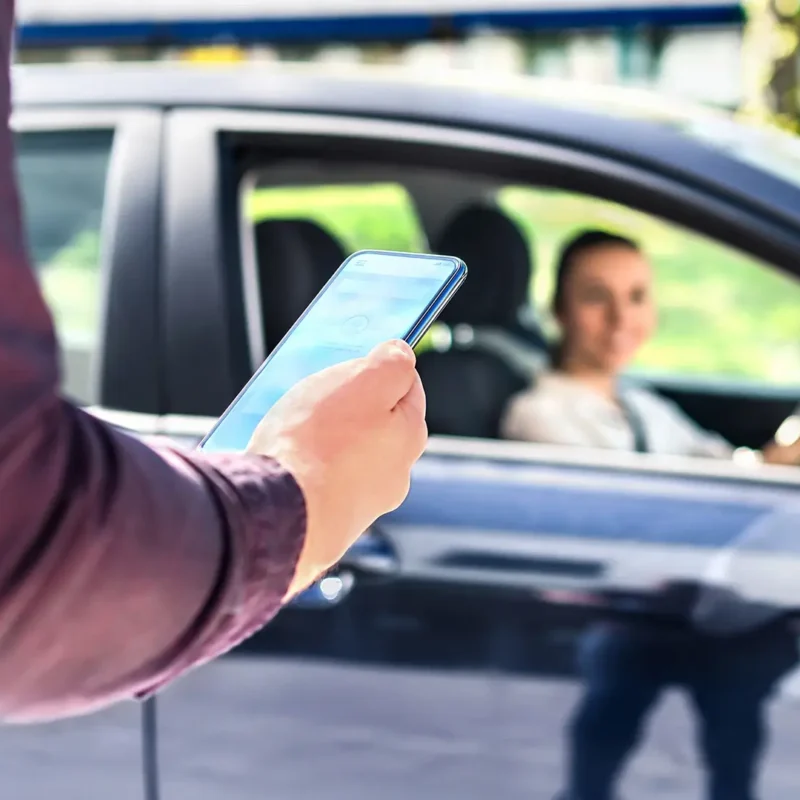 A person is holding a smartphone near a car, with a smiling female driver looking at them through the passenger window.