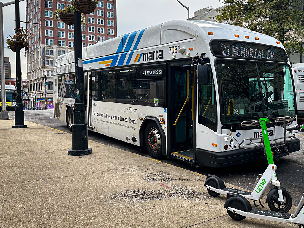 A white transit bus is stopped at the curb with its door open. There are two e-scooters nearby.