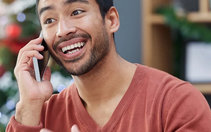 A man is sitting at a desk with an open laptop. He is holding a cell phone and smiling as he speaks. Behind him is a decorated Christmas tree.