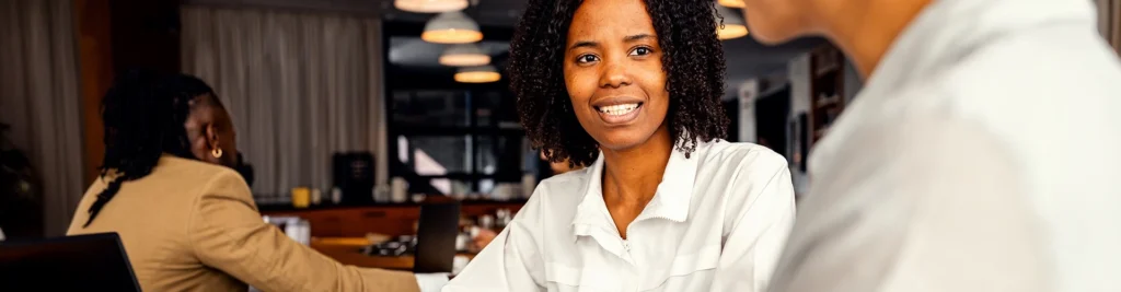 A few people are sitting in an open workspace. At the main table, a woman wearing a white shirt faces the camera and is listening to her coworker. They have reports spread out before them.