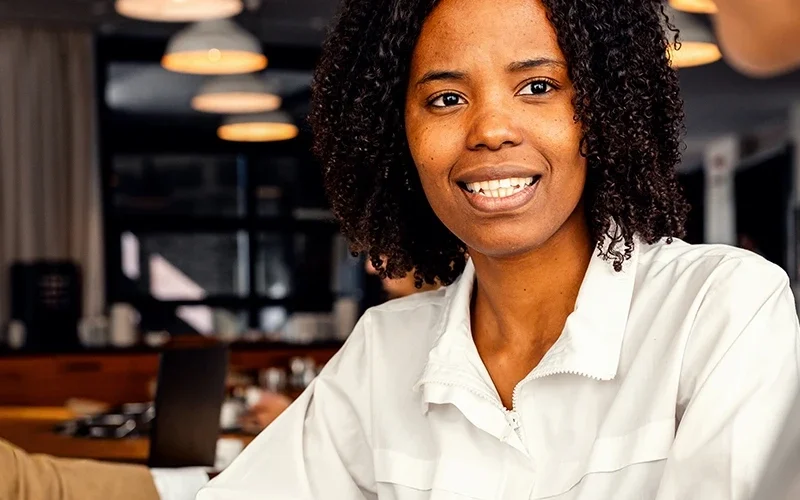 A few people are sitting in an open workspace. At the main table, a woman wearing a white shirt faces the camera and is listening to her coworker. They have reports spread out before them.